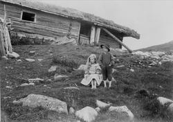 Barn i landskap foran laftet hus, ukjent sted.
Serie tatt a