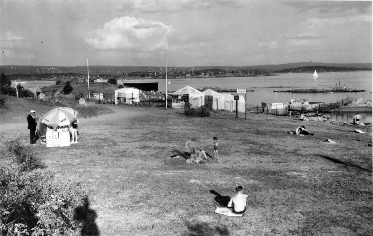 Friluftsliv. Antakelig Huk, Bygdøy. 1935. Utsikt mot øyene og byen. På stranden ses badegjester og i bakgrunnen flere salgsboder.  På fjorden er det båttrafikk.