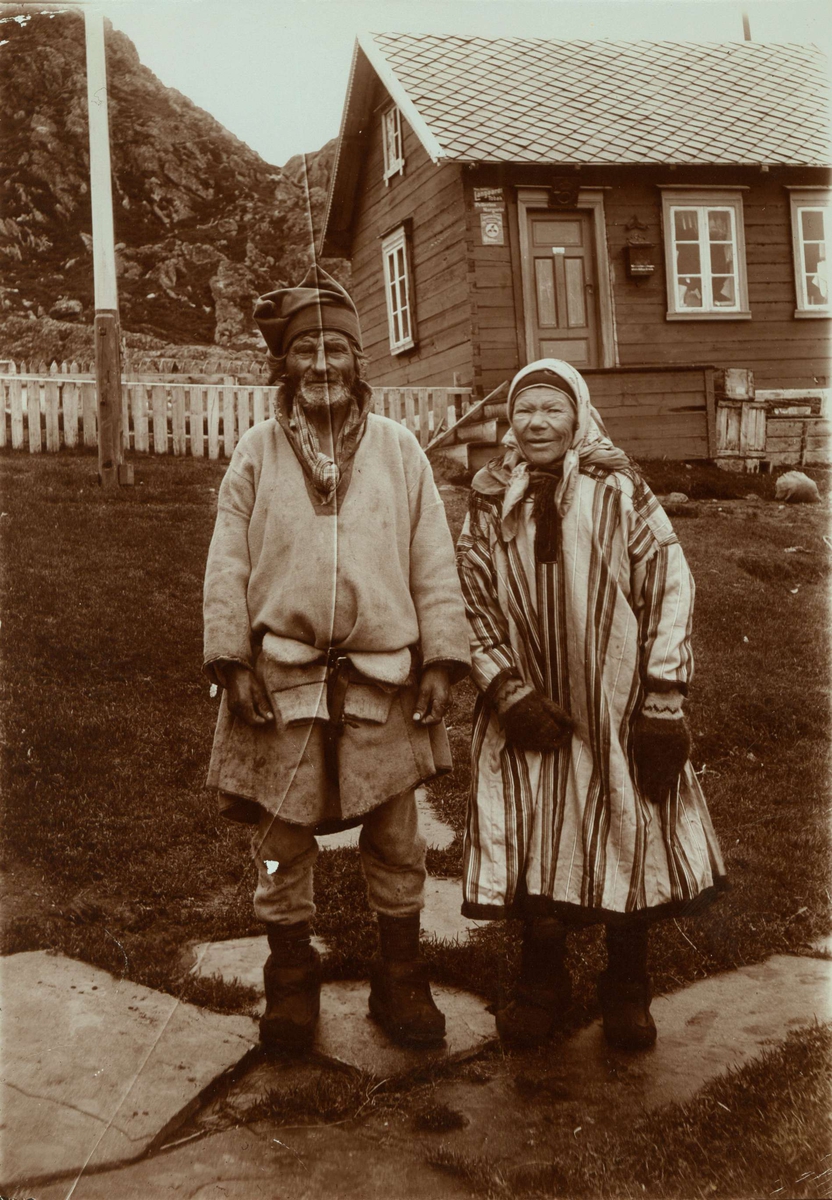 Mann og kvinne i samisk drakt, Laksefjord, Lebesby stående foran et hus med flaggstang og inngjerdet hageområde.  På huset reklameskilt for Pellerin Margarin og Langaards Tobak, muligen butikk. 
Del av serie fra en forskningsreise i Øst-Finnmark 1909.