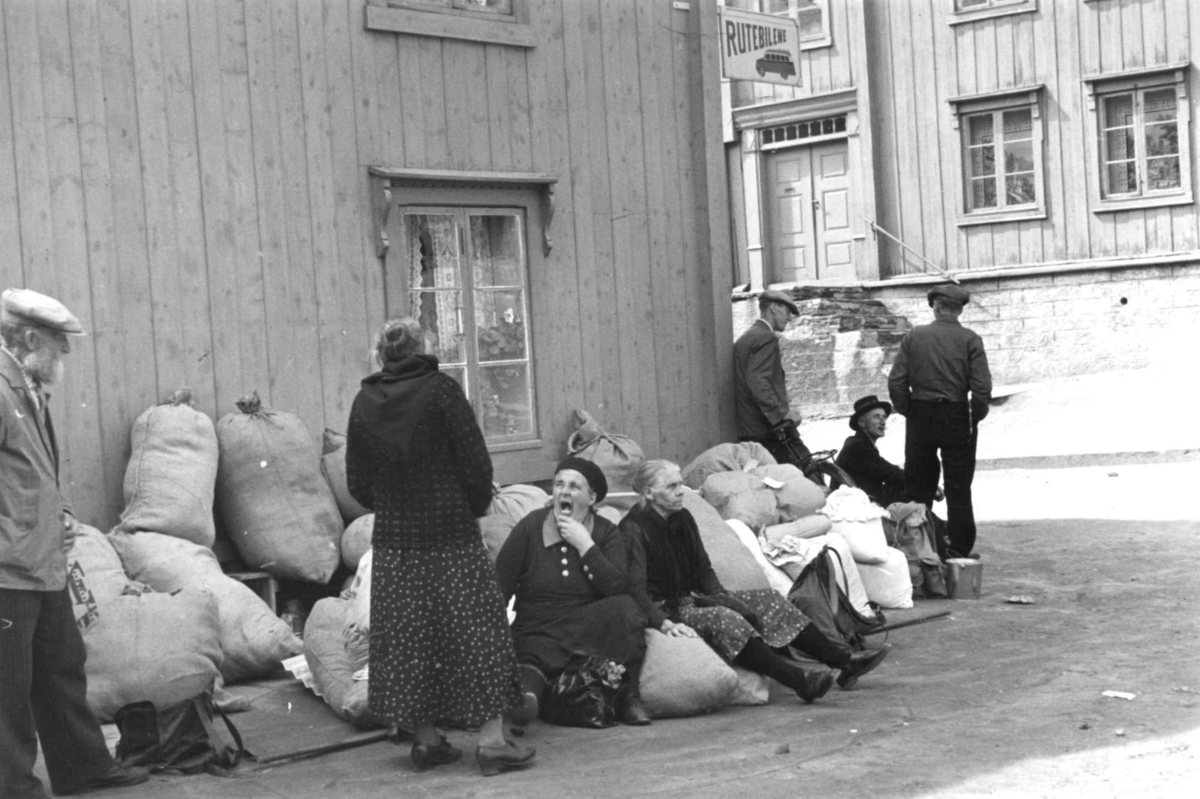 Gatebilde med menn og kvinner som står og sitter ved fulle striesekker og annen bagasje, På bygningen henger et skilt hvor det står "Rutebilene". Røros 1938.