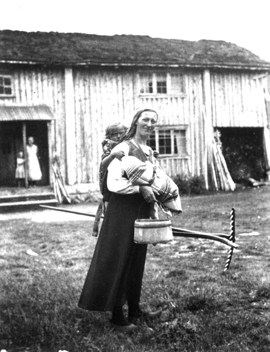 Oline Seldal med barn i fatle, i Sirdal kalt linde, et barn på ryggen og bøtte og rive i hendene, Fidjeland, Sirdal,  Vest-Agder 1936. Rekonstruksjon. Barnet hun har på ryggen er hennes niese, Tordis Harbo, f. Fidjeland (f. 1932).Et hus i bagrunnen med kvinne og barn i inngangspartiet. Oline Seldal var bosatt i Høle, Sandnes, Rogaland.