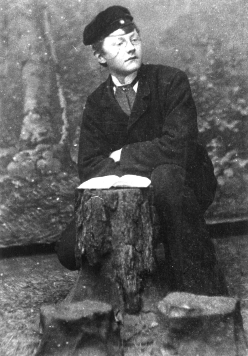 Ung mann sitter ved en stubbe med en bok på, antagelig i et fotostudio. Anna Grostøls familie. Vest-Agder 1941-42.