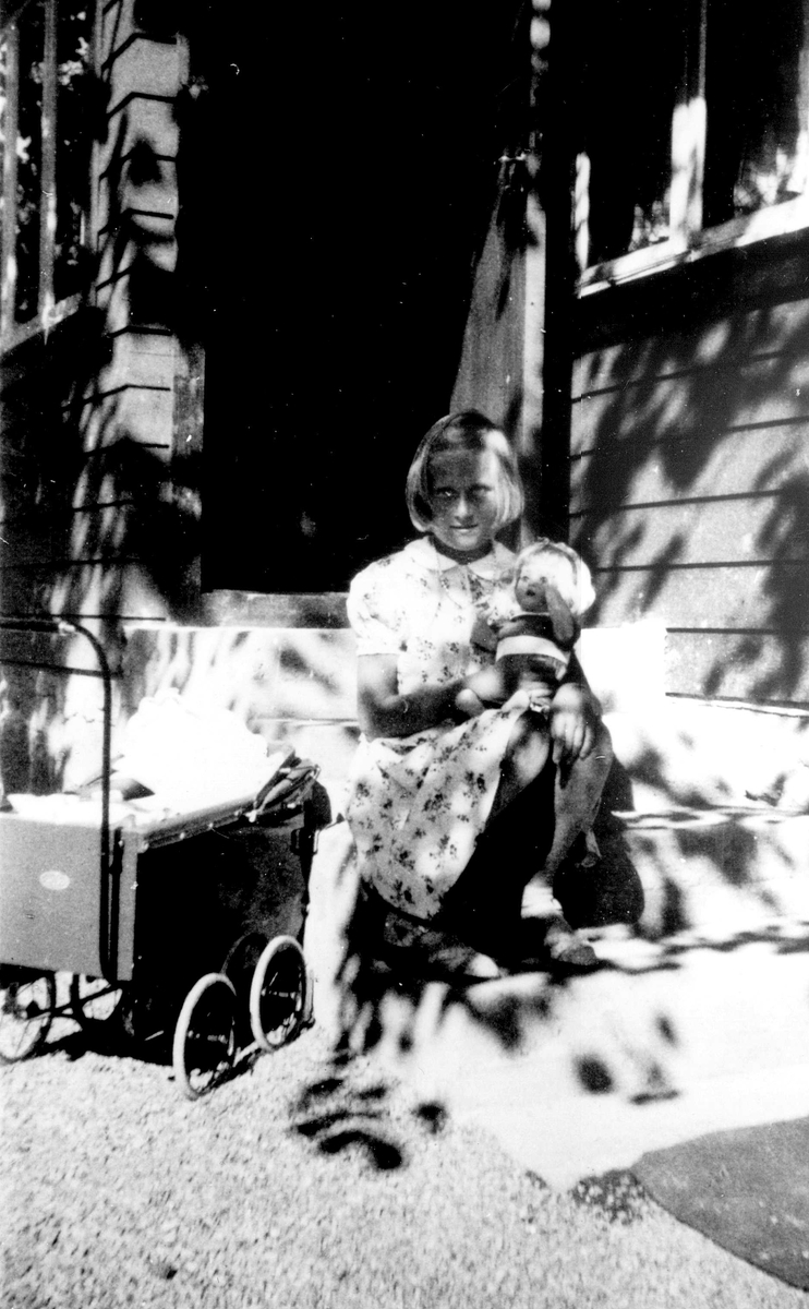 Jorunn Fossberg med dukker og dukkevogn foran sommerhus. Bildet er tatt ved familien Fossbergs sommerhus på Nærsnes i Røyken, Buskerud som ble tatt i bruk sommeren 1937. 
Fra Jorunn Fossbergs familiealbum.