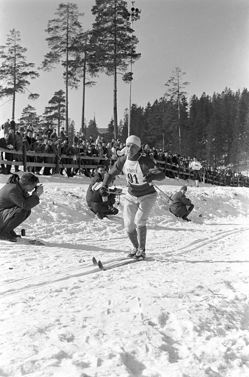 Skiløper med startnummer 91 i Holmenkollen. Publikum langs løypa.  Pressefotografer på sidelinjen. Holmenkollrennene 1963.