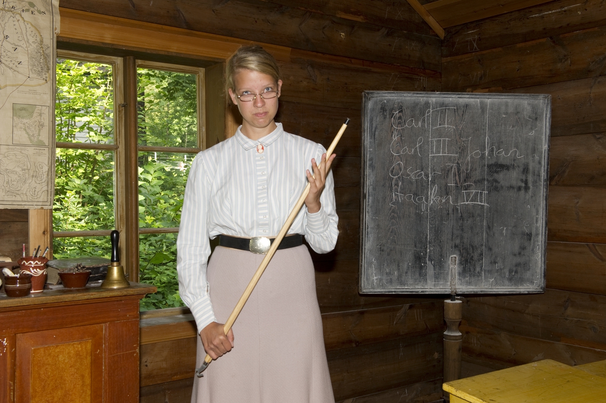 På historisk ferieskole får barn kjennskap til tradisjoner og historie gjennom opplevelser, lek og læring. Her står frøken Åse i skolestua med pekestokk.