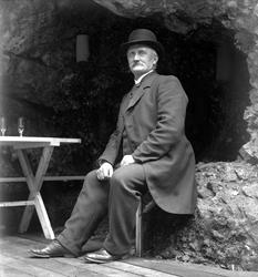 Mann sittende på benk på brygge med berget i ryggen. Avfotog