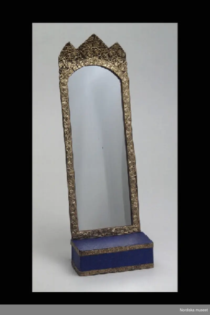 Inventering Sesam 1996-1999:
a) spegel  H 26,5   B 9,5  (cm)
b) bord  L 10   B 4,5   H 3,5  (cm)
Trymå, spegel med bord, av papper, till dockskåp. Hög, rektangulär spegel, ram av präglat guldpapper, uppdraget krön som delas i tre spetsar. Lådformat bord av blått papper med guldfriser
Tillhör dockskåp från omkr 1840, inv nr 145.505, inrett 1872 av leksakshandlare W A Bandel, Västerlånggatan, Stockholm, för dottern Thyra, vilken vid överlämnandet till museet själv gjort uppställningen av föremålen i dockskåpet.
Birgitta Martinius 1997
