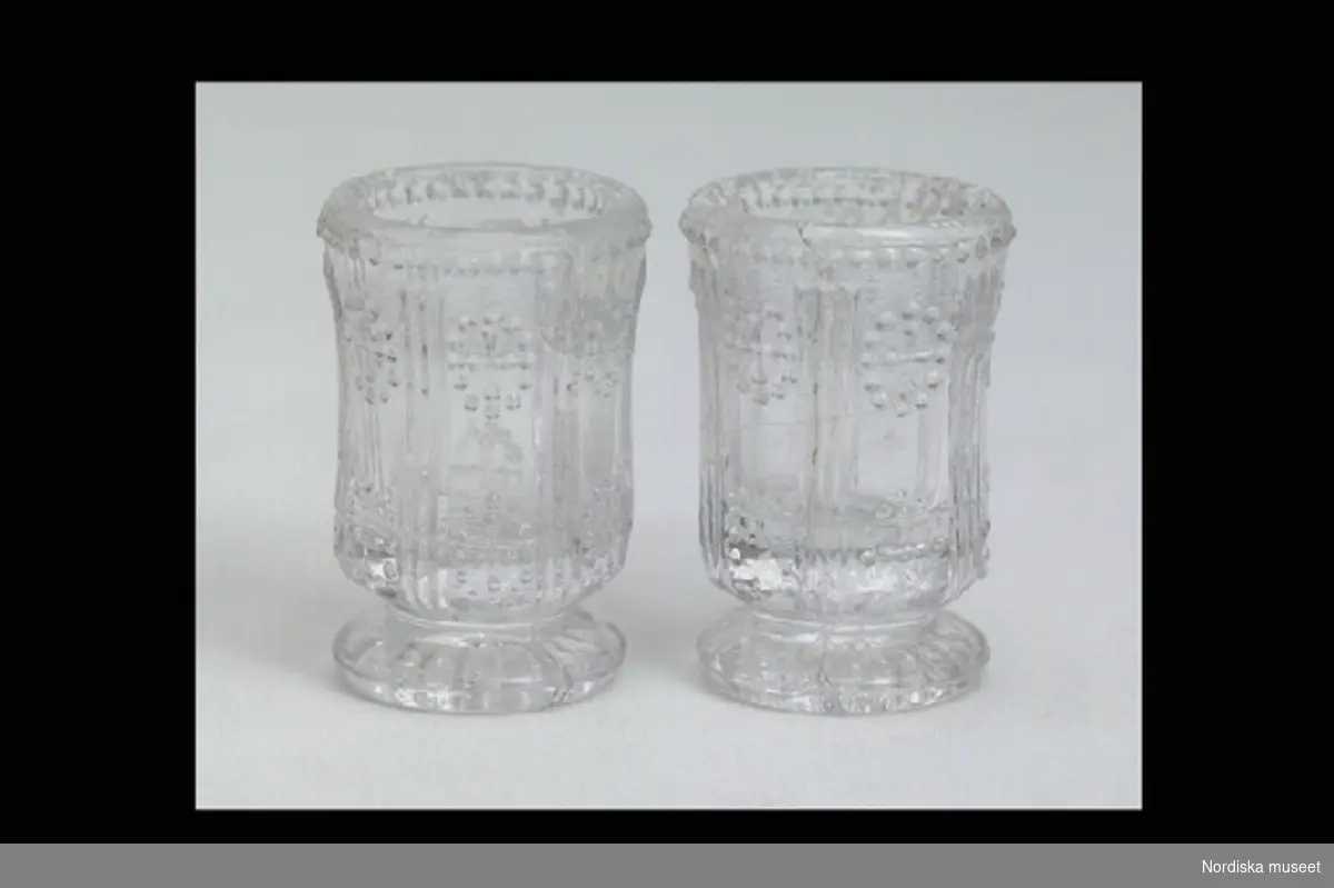 Inventering Sesam 1996-1999:
H 3,5 cm
2 st glas av pressglas med lätt utsvängd kuppa på låg fot, dekor av ränder och prickar.
Tillhör dockskåp inv. 200.513.
Birgitta Martinius 1996