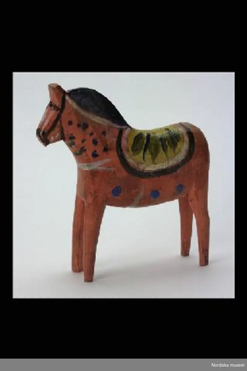 Inventering Sesam 1996-1999:
L 16,5 H 17,5 B 4 (cm)
Häst av trä, rödmålad med krusning i gult, blått, svart och vitt. 
Delvis bestruken med gulnad fernissa. 
Birgitta Martinius 1996