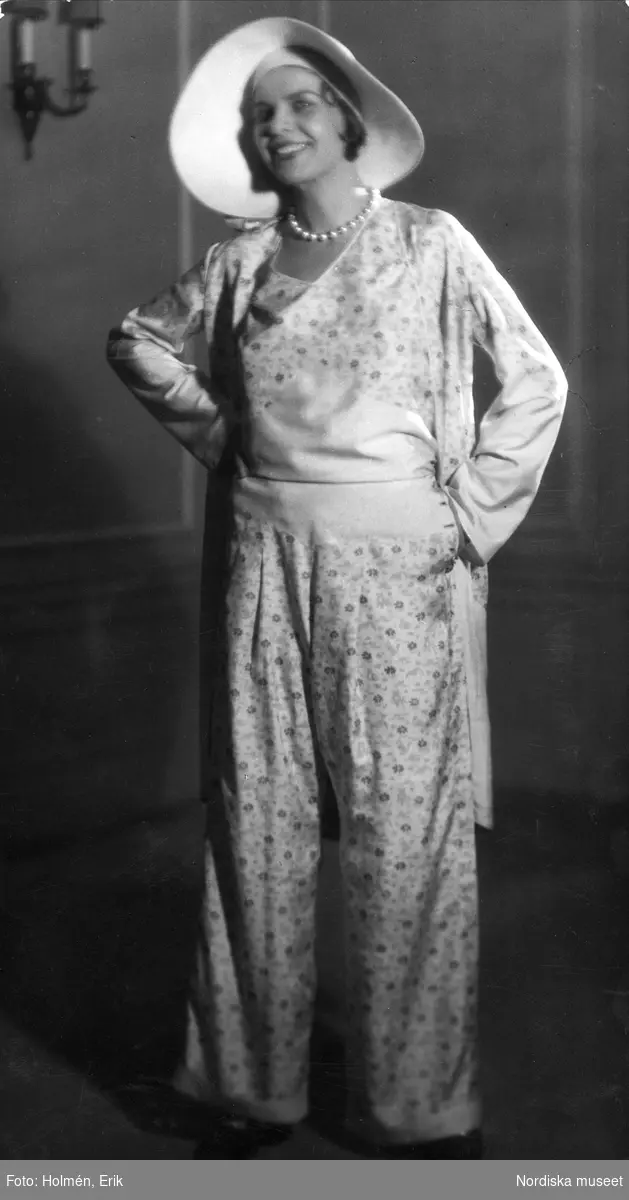 Modebild från Nordiska Kompaniet. Jean Patou. Modell klädd i vida byxor och lång överdel med småblommigt motiv, samt vit hatt och vitt pärlhalsband.