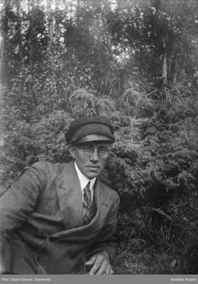Edvard Olsson i kostym, vegamössa och glasögon ute i skog och mark. Självporträtt.