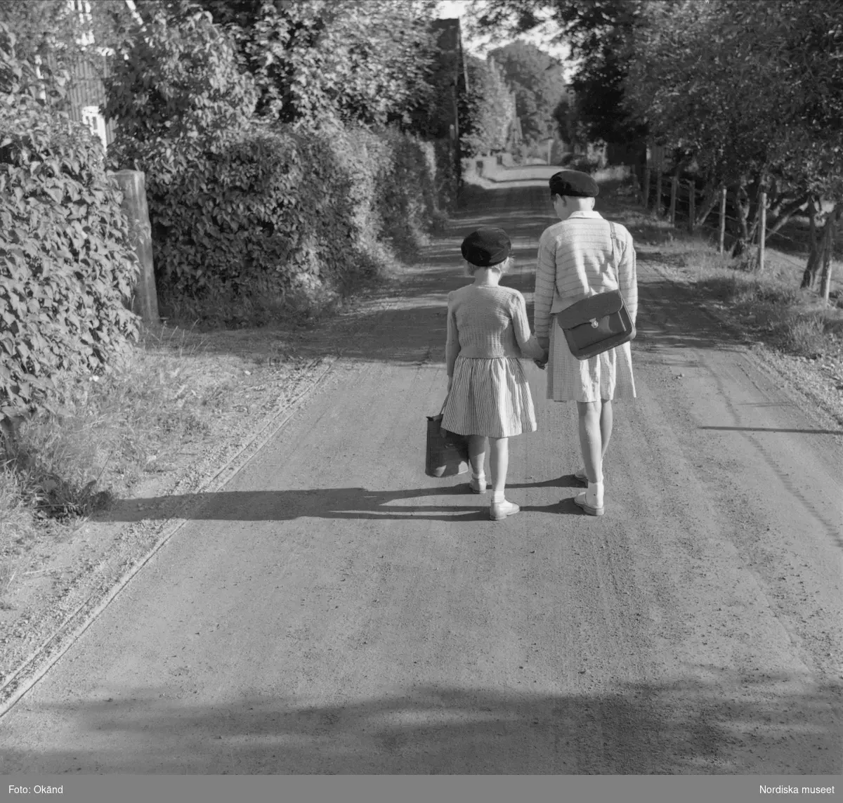 Bild ur frågelistsvar. Två flickor promenerar i solskenet på landsväg med ryggarna mot fotografen. De håller varandra i handen och bär på väskor.