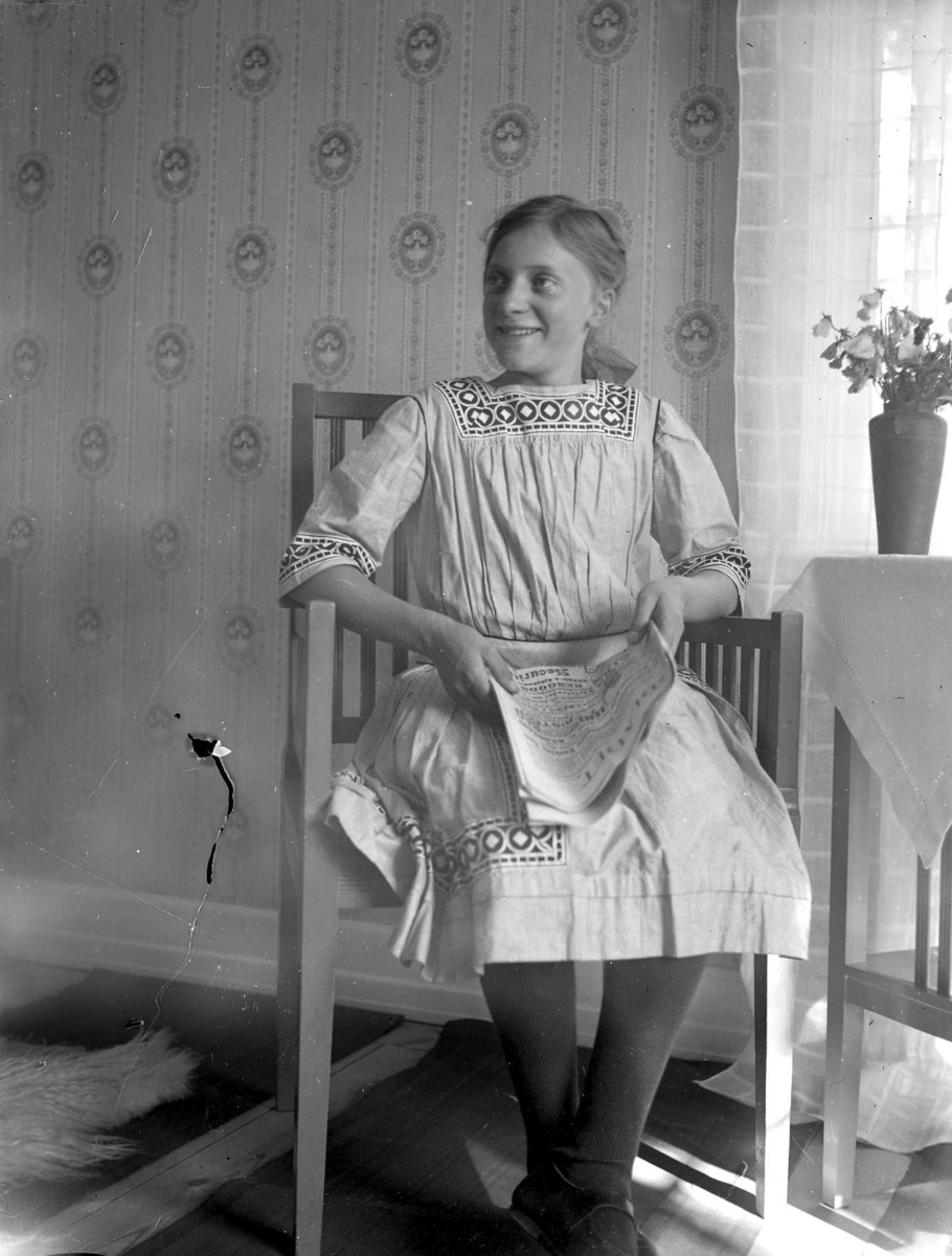 En flicka sitter i en stol vid ett bord. I knät har hon en tidning, på bordet står en bukett luktärter i vas.