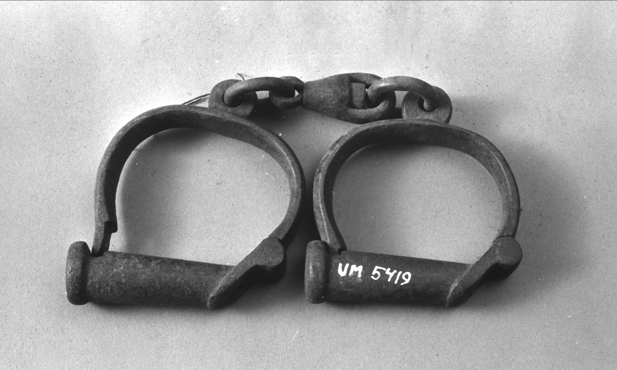 Ett par handbojor av järn sammanfogade med en järnlänk. Handklovarna är utformade som hänglås.
