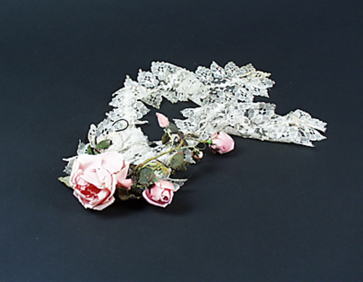 Hårklädsel av vit tyllspets. Prydd med en ros och sju rosenknoppar samt ett guldfärgat fröhus och gröna blad. Stomme av ståltråd och svart tyll.

