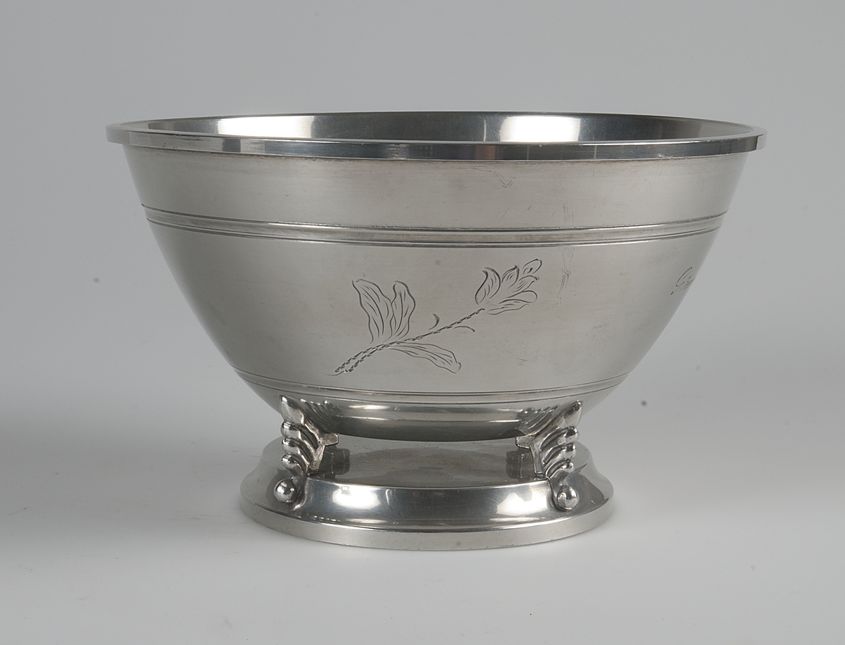 Tennskål med fotplatta. Skålen har fyra dekorativa "fötter" som  håller samman skålen med fotplattan. Skålen är vid och har ingraverade blommor och texten: Grand Prize 1933.