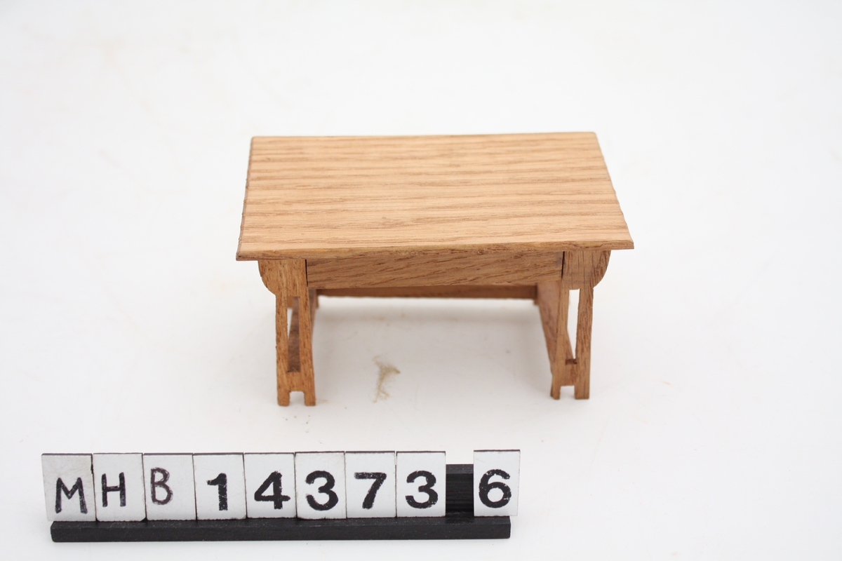 Miniatyrbord til dukkehus. Det er i tre, og har bordplate med rektangulær form. I hvert hjørne er doble bordbein som nede er forbundet med hverandre både i lengde- og bredderetning.