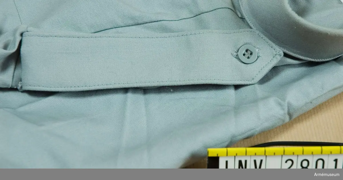 Dubbelvikt med smalare underdel. Knapphål på båda delarna för fastknäppning i den hälla som finns på skjortan. Fastsättning i ärmsömmen.