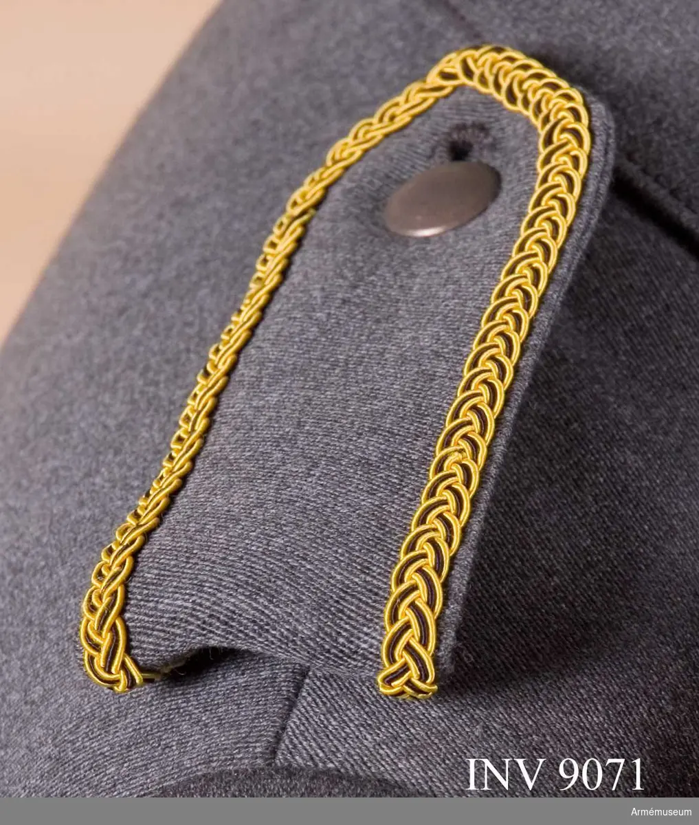 Av samma tyg och färg som jackan kantad med en flätad galon i gult och svart silke. Tecken för kårchef. Axelklaffen är dubbel och viks runt en träns vid axelsömnen och knäpps mot en grå knapp av mindre modellen på jackan.