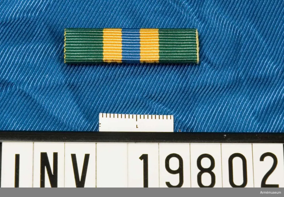 Grönt band med en blå rand i mitten åtföljd av en gul rand på vardera sidan. Släpspännet förvaras i ask tillsammans med en medalj och en miniatyrmedalj.