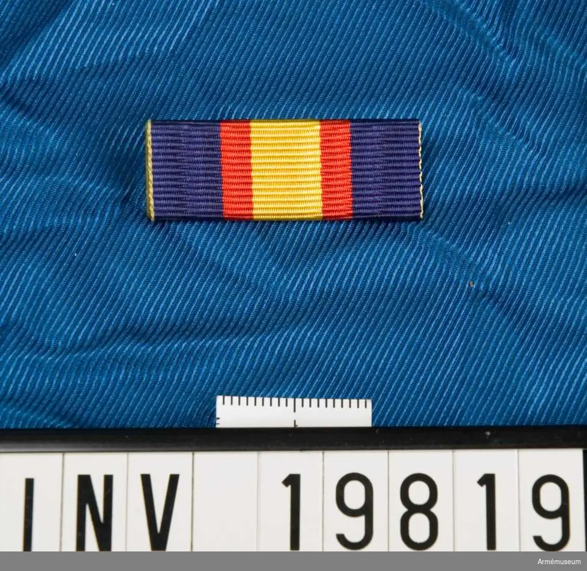 Blått band med en bred gul rand på mitten åtföljd av en röd rand på vardera sidan. Släpspännet förvaras i en ask tillsammans med en medalj och en miniatyrmedalj.