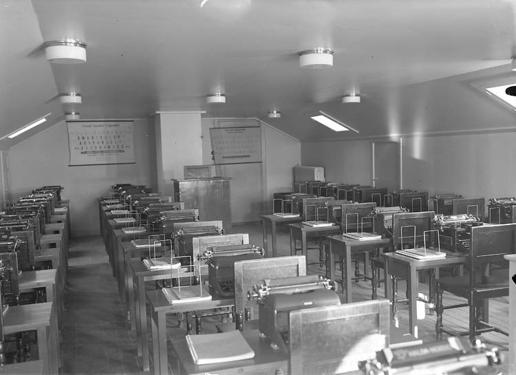 Text till bilden:"Skolsal med bord och skrivmaskiner i fyra lång rader".