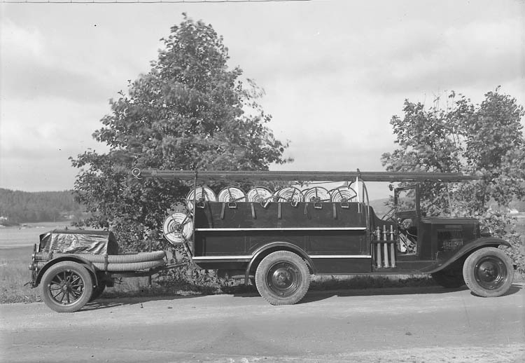 Text till bilden:"Brandbil på en grusväg med stegar, slangar och en släpvagn. Ljungskiles första brandbil".