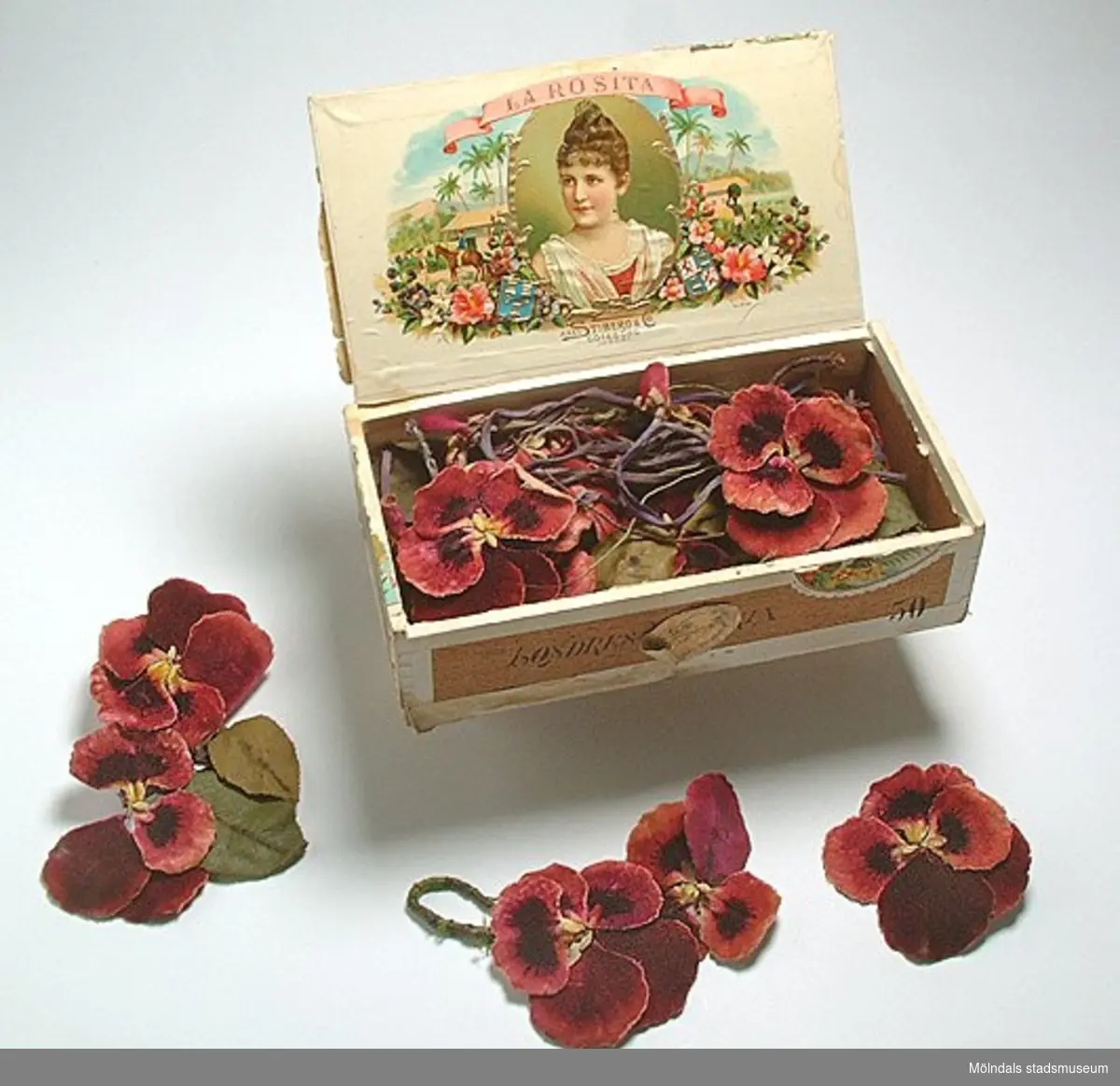 Blomgarneringar i form av vinröda penséer med sammetsyta. Förvaras i en cigarrlåda.