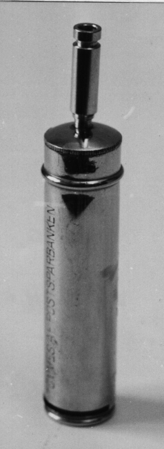 Ficksparbössa i förnicklad mässing, i form av en
cylinder.Låsenhet upptill i locket som skruvas av med en skruvnyckel,
locket vänstergängat. På övre halvan en gördel runt sparbössan.
Längscylindern finns hål med siffror bredvid, detta för att kunna se
hur mycket pengar som fanns i sparbössan. Denna första typ av
ficksparbössa infördes 1906, och de kunde köpas för 1 krona/st. Från
19220101 är priset 75 öre/st. Denna typ ersattes år 1926 av en
ny.Försäljningen upphör 1967, då ytterligare en typ, i plast, tas i
bruk. Sparbössan har plats för 5 kronor i 10-öringar, och var avsedd
att bäras med i fickan, för att stoppa växelmynten i, och därmed
underlätta sparandet. Med nyckel.