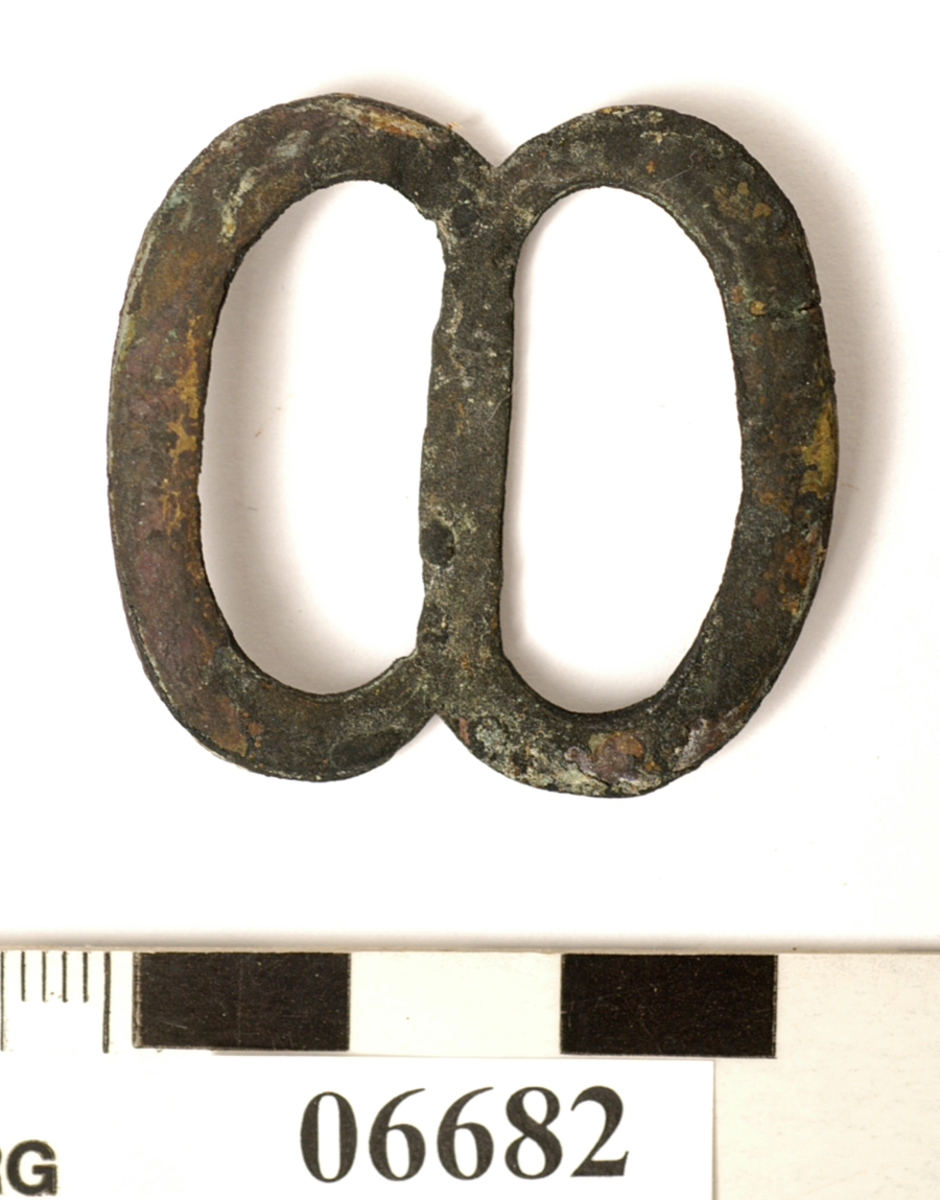 Sölja. Spänne. Formad som en åtta. Lätt böjd på mitten.

Text in English: One metal buckle.
One side of metal (probably front) is convex, the other side (probably back) is flat, with defined edges.
The whole piece is concave, the center bar sits lower than the sides.  When viewed from the side, it resembles a wide V.  When viewed from the front or the back, it resembles a flattened figure 8.
Handmade out of what appears to be one piece of metal, two solder points visible.