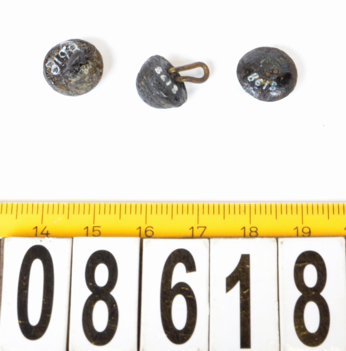 Skoknappar, 3 stycken av mässing.
2 med ögla, 1 utan. Ornering på knapphuvud (varierande).

Text in English: Three decorated metal buttons.  Two with similar but different patterns.  All are half-sphere shape with almost flat bottoms.  Two have wire shanks, one is missing.
First pattern is a lattice.  Six lines are woven together and each space between is filled with five dots. This button has a wire shank that is cast in far offcenter.
The second button has a flower at the top in the center with a circle around it.  Under this is a pattern that is too worn to be accurately described, but appears to be a four-repeat swoop pattern.  The wire shank is cast into the head, and is severely bent to one side.
The third button has a five-petal flower in the center at the top with a circle around it.  The pattern below it is a five-repeat pattern of opposing "C"s, connected by a line in the center and with dots above and below the line.  There is no shank, but holes in the bottom indicate there was probably a wire shank at one time.