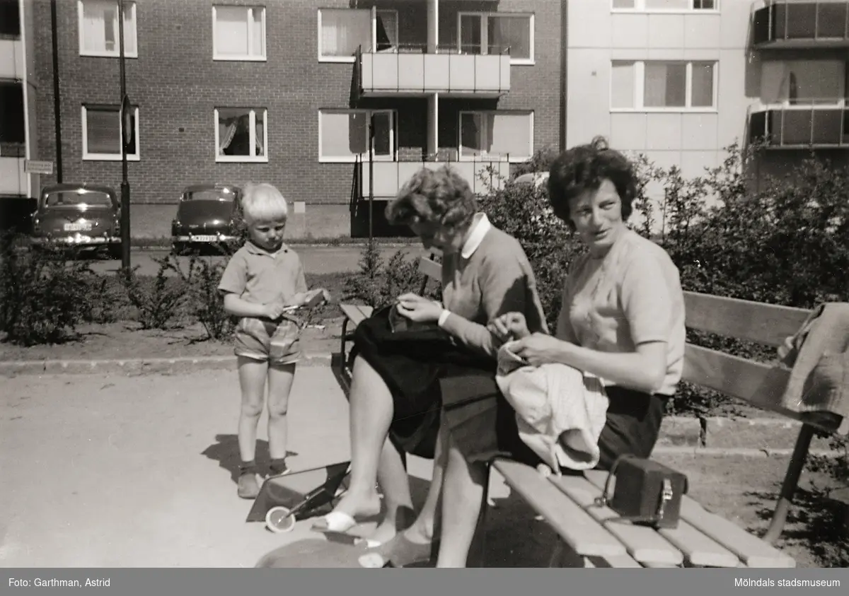 Två kvinnor, Lisbert Andersson och Kyllan Lageklöv, sitter på en bänk på Lerbrännaregatans innegård, 1960-tal. De passar på att handarbeta när barnen leker. Till vänster står ett barn. Kvinnorna är bekanta till fotografen Astrid. Familjen Garthman flyttade till Lerbrännargatan 1C, 1960-09-01.