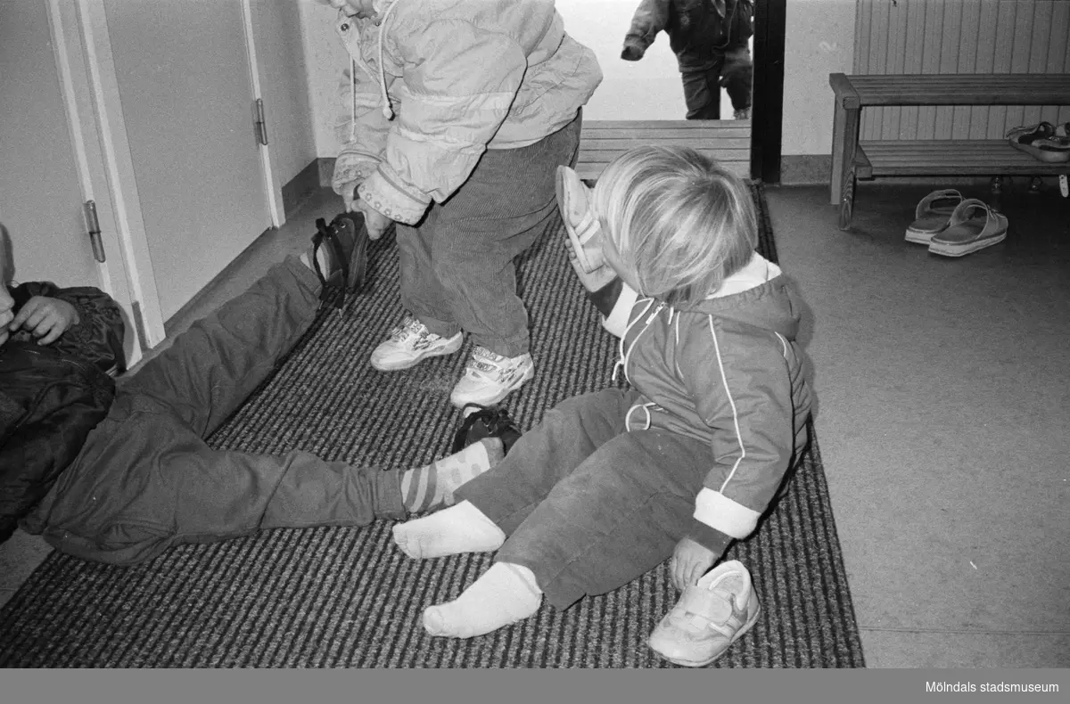 Tre små barn sitter/står på entrémattan och hjälps åt med av-/påklädning av ytterkläder. Katrinebergs daghem.