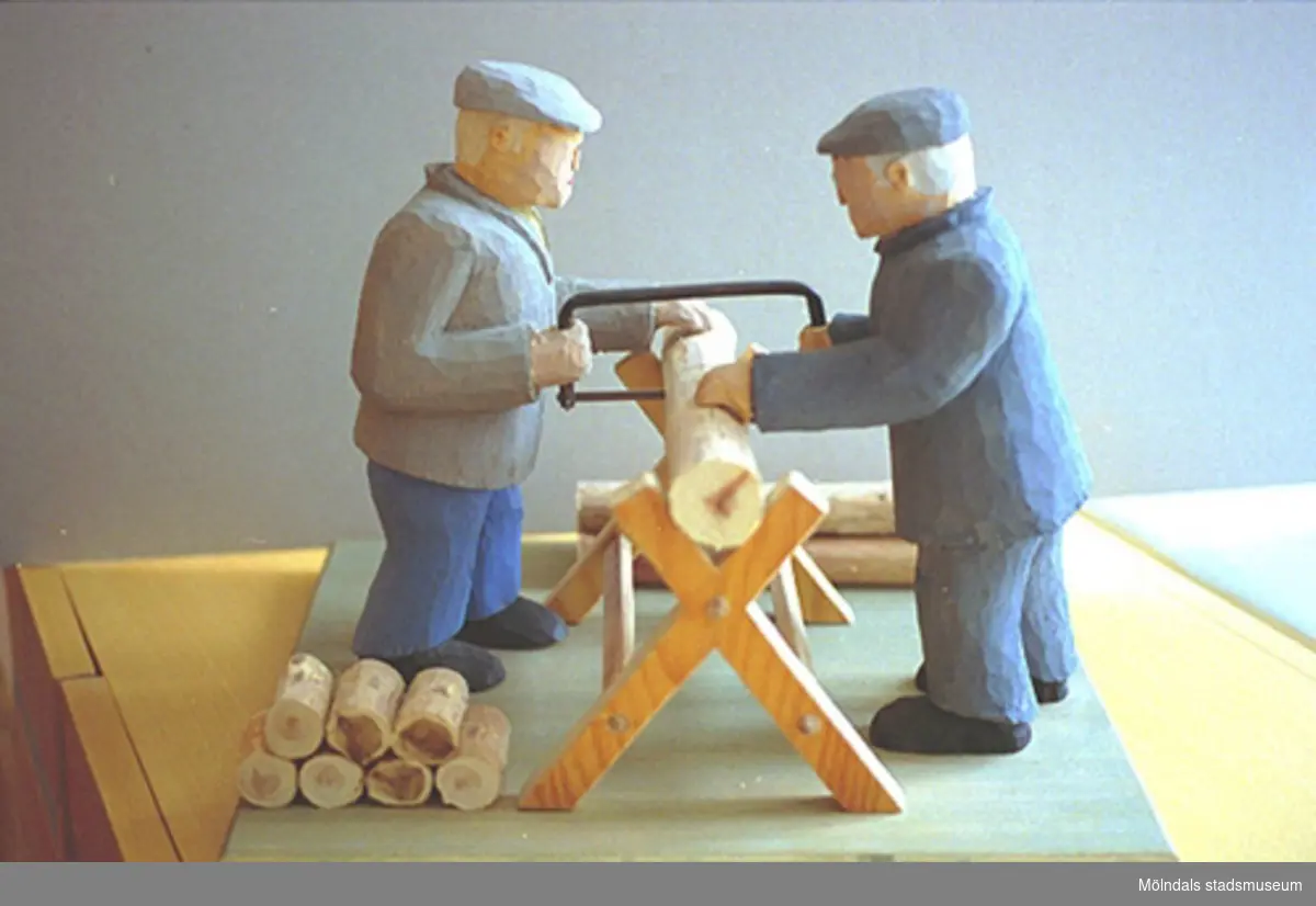 Två män står på var sin sida av en sågbock. De hjälps åt att såga ved. Harry Bergmans "gubbar" (träfigurer).