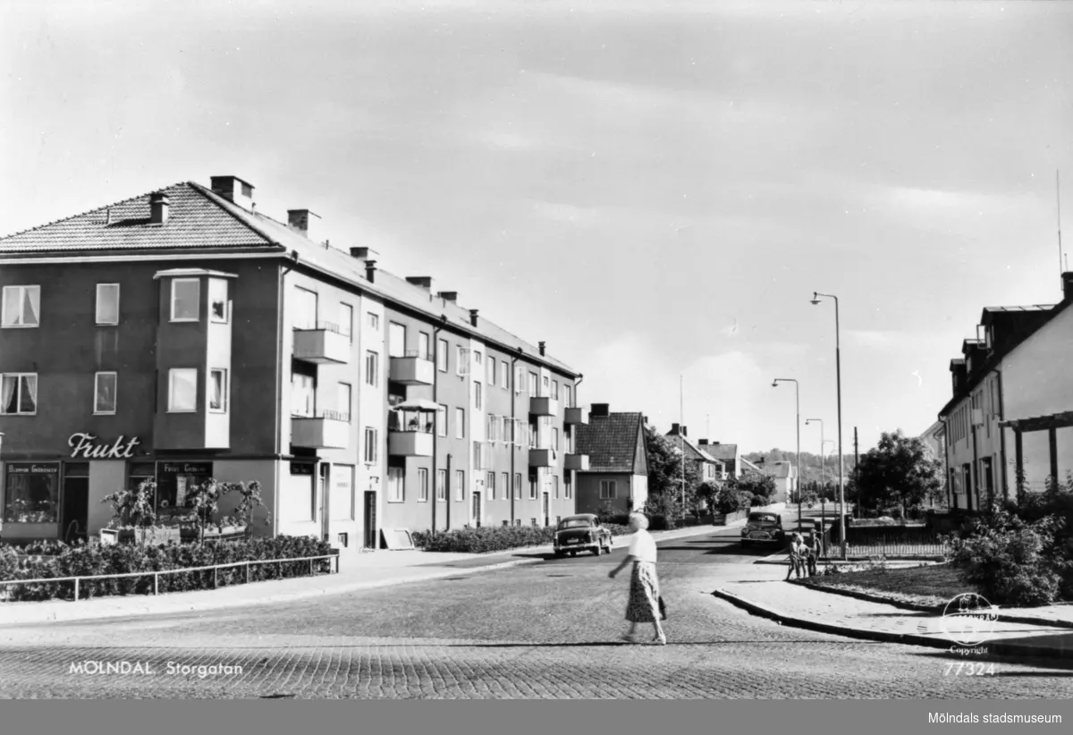 Vykort "Mölndal. Storgatan" från 1950-tal. Utsikt från Frölundagatan utefter Storgatan söderut. Till vänster (husets gavel) ligger Brogatan 23 (Berghedes Frukt & Blommor, Danskens bageri på hörnet, senare Järnhuset). Lägenheterna ligger på Storgatan 33A-C. Till höger skymtar Storgatan 34-36.