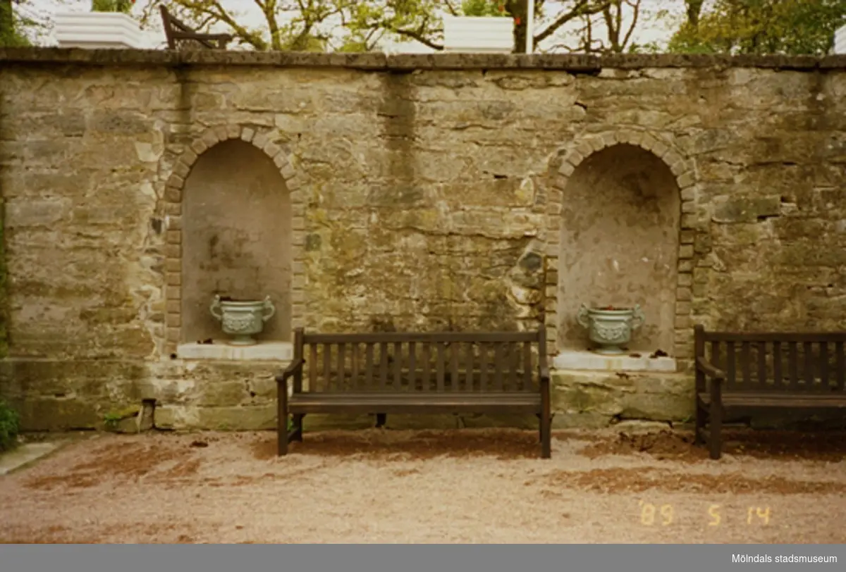 Två parksoffor står framför en stenmur (kejsarterrassen). På muren samt i de två nischerna står det två blomkrukor.