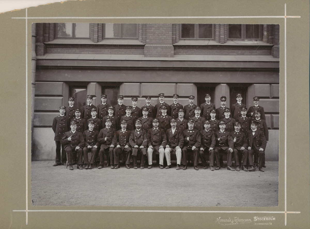 Fotografi föreställande samtliga brevbärare, rekbrevbärare och brevbärarförmän vid Postkontoret Stockholm 4, ca 1910. Fotot är taget utanför Högre Allmänna Läroverket.