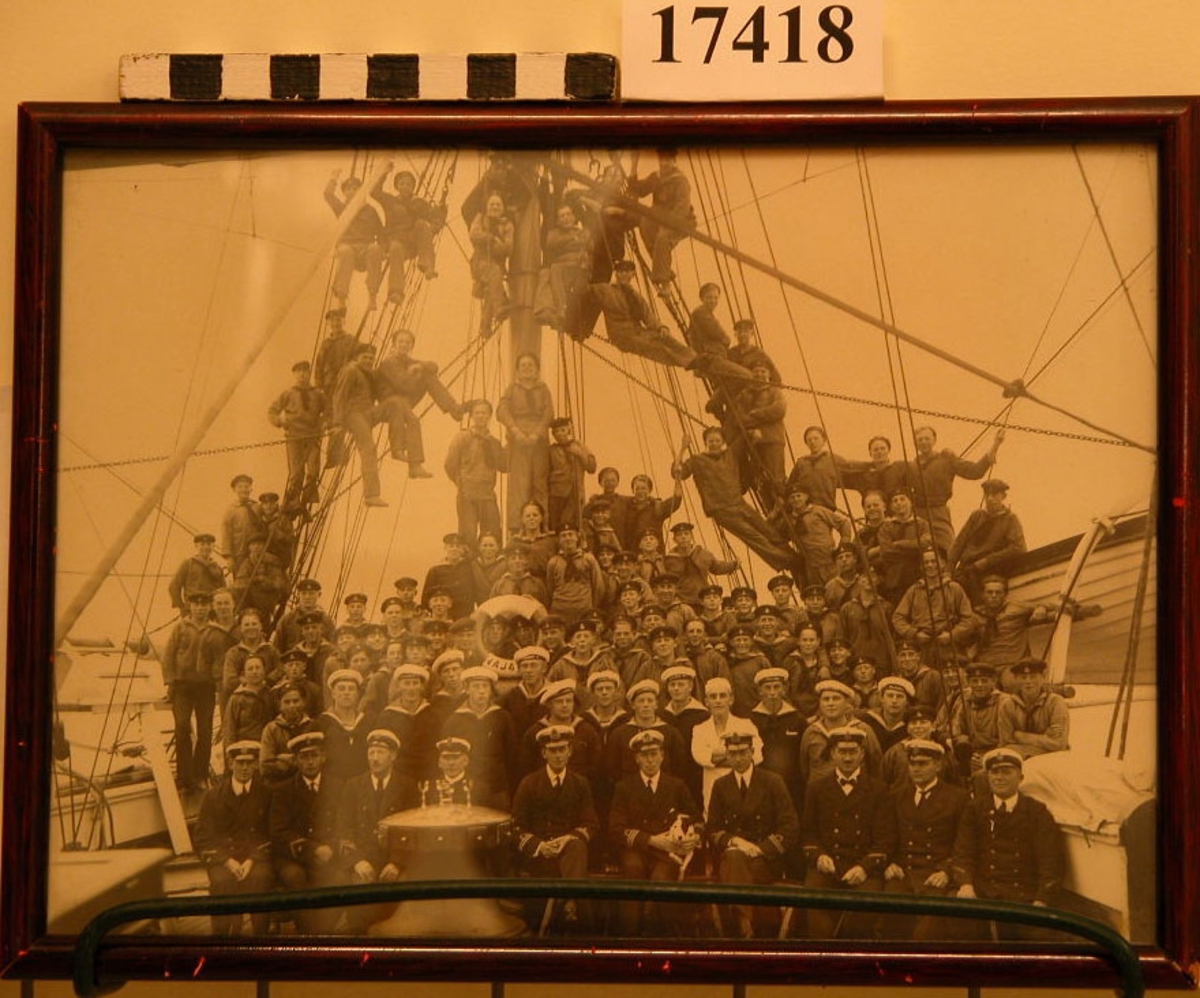 Fotografi inom glas och ram visande besättningen på HMS Najaden stående på däck och klättrande i riggen. Befälen sitter längst fram, skeppsgossarna är placerade bakom. Ramen är av trä, mahognyfärgad, B= 9 mm. Baksidan består av papp med handskriven text: "HM "Najadens" Besättning sommaren 1923" samt givarens namn.
.