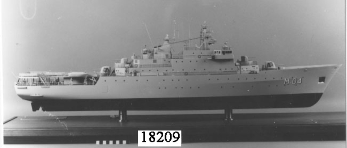 Fartygsmodell av metall och trä föreställande minfartyget Carlskrona placerad i monter. Målad i ljus grått, svart under kölvattenlinjen. Två propellrar i mässing och två roder. Skråvet har två rader ventiler, i fören målat med vitt " M 01 ". Däcket är mörkgrått. Livbåtar, i aktern helikopterplattform märkt med stort orange H. Luftvärnspjäser i för och akter, antenner. Modellen är placerad i monter bestående av rektangulär teakplatta med glashuva och är fästad till plattan med hjäp av två hållare av mässing. På plattan finns två metallplattor med graverad text placerad på vardera kortsida, samma text på båda plattorna. "Minfartyget Carlskrona, leverantör Karlskronavarvet AB, leveransår 1982, Byggt för försvarets materielverk, Längd överallt 105,70 m, Bredd mallad 15,20 m, Djupgåend mallat till KVL 4,00 m, Deplacement till KVL inkl. bihang och bordl. 3138 kubikmeter, Maskinstyrka 4 x 1940 k W."
Monterns mått: H = 420 mm  L = 1170 mm B = 260 mm