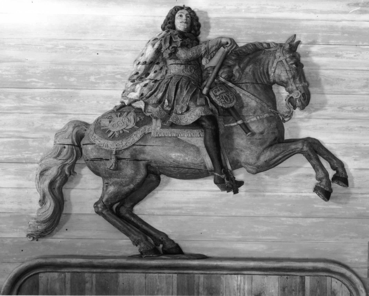 Akerspegelornament, ryttarfigur, föreställande Carl XI till häst. Nära naturlig storlek. Hästens vänstra sida avskuren till plan yta för anliggning mot akterspegeln. Konungen med kommandostav, kappa och pistol i hölster. Stigbyglar och betsel av järn allt övrigt skulpterat trä. Häst mörkgrå, konungen i ljusbrunt, rött, blågrönt och svart.

Suttit på akterspegeln till linjeskeppet Sverige, tidigare kallat Carolus XI, byggt 1677–79 i Stockholm.