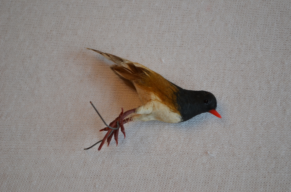 Fugl av papp, dekorert med fjær. Bein og nebb er av plast. Ståltråd på beina slik at den kan festast i eit juletre.