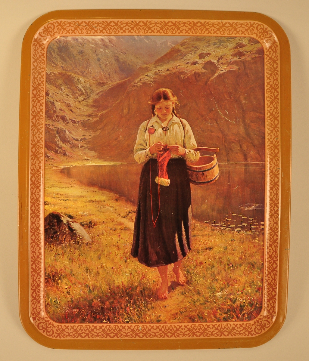 Nasjonalromantisk motiv av ei jente med fletta hår som går berrføtt i graset ved eit vatn og strikkar. På armen ber ho ei trebøtte. I bakgrunnen eit fjellandskap.