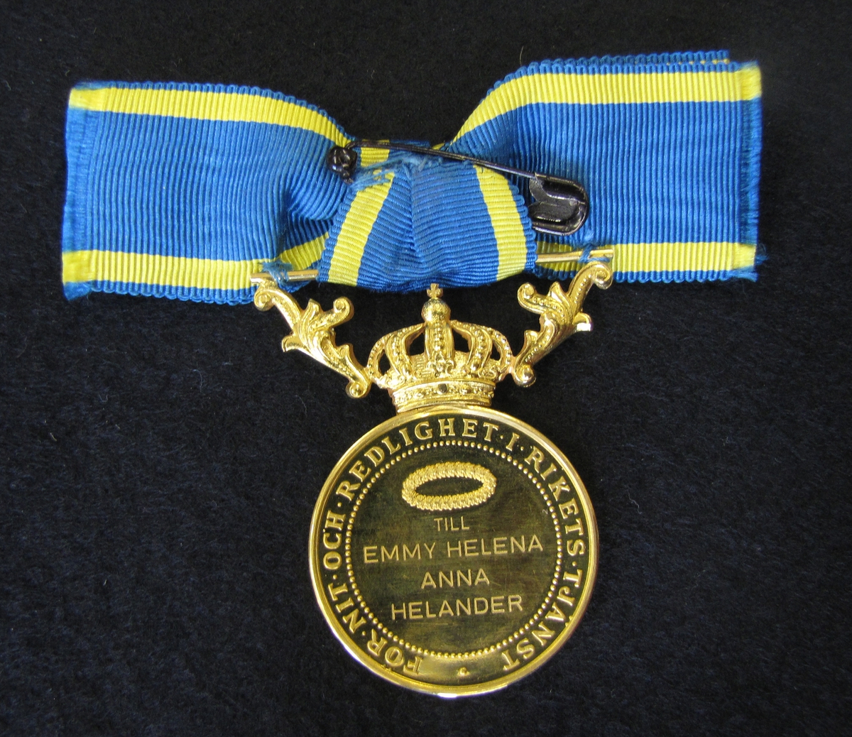Medalj till Emmy Helena Anna Helander för nit och redlighet i rikets tjänst.  Finns ett etui till medaljen (VM09633:c). 

Medaljen är av 23 K guld försedd med krona och bärornament av 18 K guld.