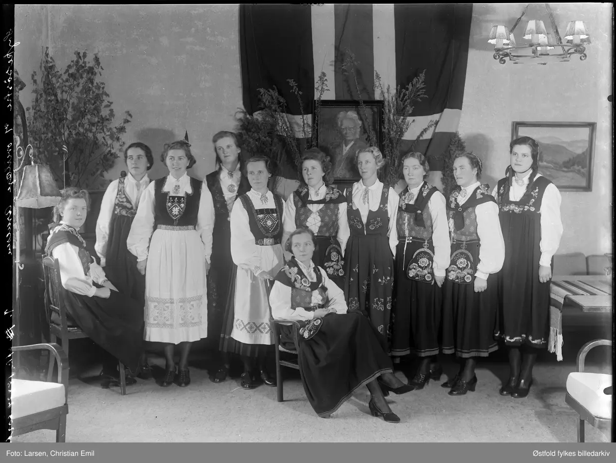 Gruppe kvinner kledd i forskjellige bunader. I bakgrunnen flagg og portrett av Henrik Ibsen. Pyntet med greiner, jubileum?
Sykesøstre med kona til overlege Arne Arnesen sittende foran?? Eller er kvinna overlege Arnesen?