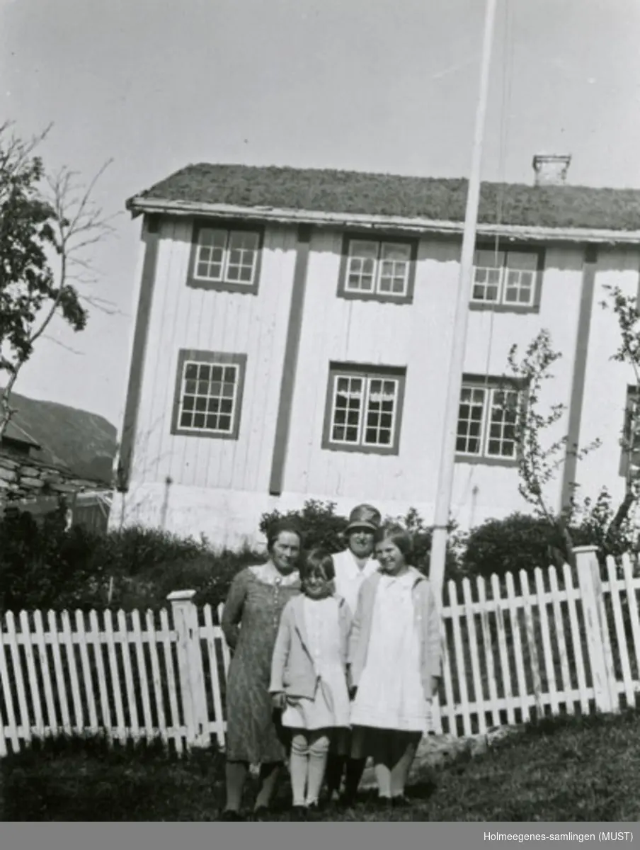 To kvinner og to jenter i sommerkjoler står foran et hvitmalt tregjerde, i bakgrunnen et bolighus med to etasjer. Se også ST.K.HE 2007-011-0111 til -0126.