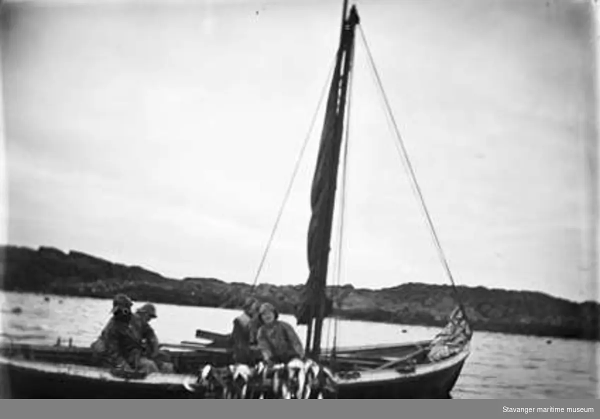 Fem fiskere som trekker sildegarn. De er ombord i en robåt med beslått seil.
