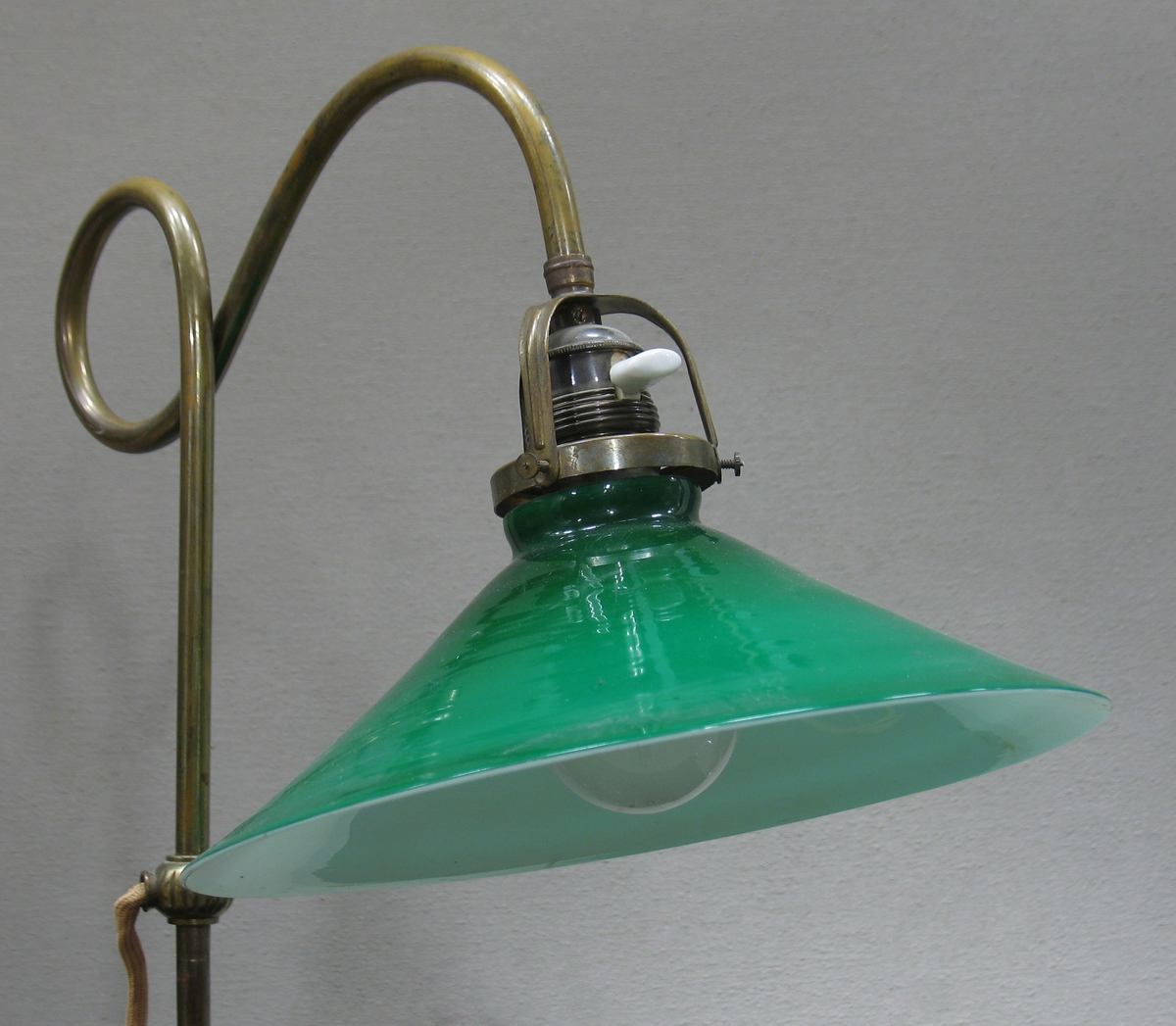 Skrivbordslampa med fot i mässing och grön glaskupa. Både fot och ställning är fint dekorerade och från mitten av ställningen löper en sladd som avslutas med stickpropp. En glödlampa är fäst innanför kupan.