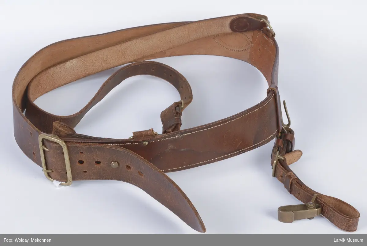 Form: Bredt belte med spenne for feste. Et smalere belte som går over skulderen.  Det brede beltet har en krok, og en stropp som henger ned.
