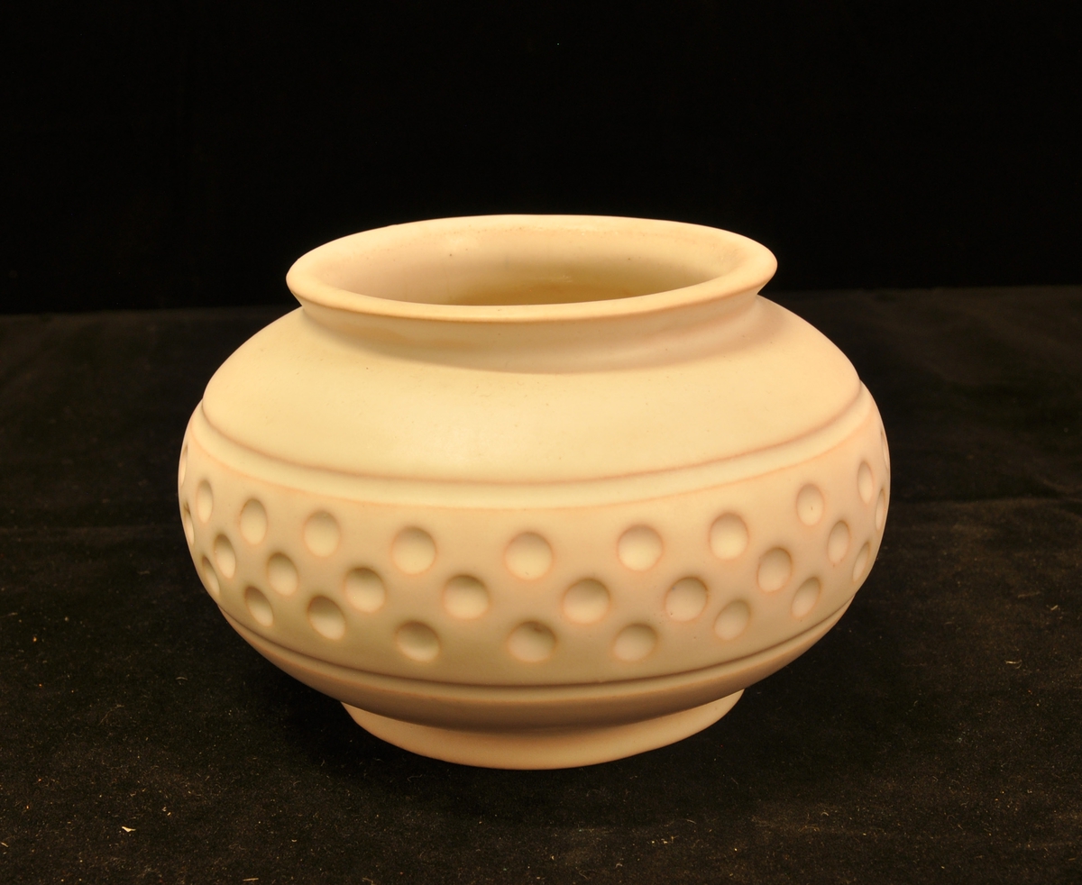 Vase i keramikk, lav og vid med kraftig utbuking. Midt på en bord markert med linje oppe og nede og små runde fordypninger. Ensfarget utvendig med tynn grå glasur. Det rødlige godset skinner delvis gjennom.
Modellnummer R. 5161A 