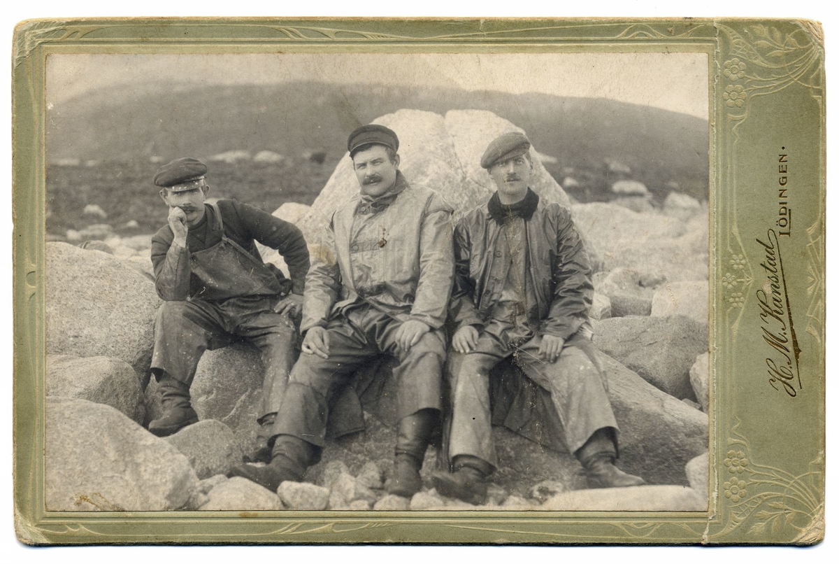 Gruppeportrett av tre menn i arbeidsklær, fotografert utendørs foran en stor stein. Bildet skal være tatt i forbindelse med sildefiske.